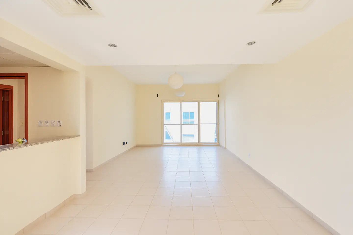 Empty, bright room with beige walls and tile floor. A sliding glass door leads to a balcony. A kitchen counter is visible on the left.