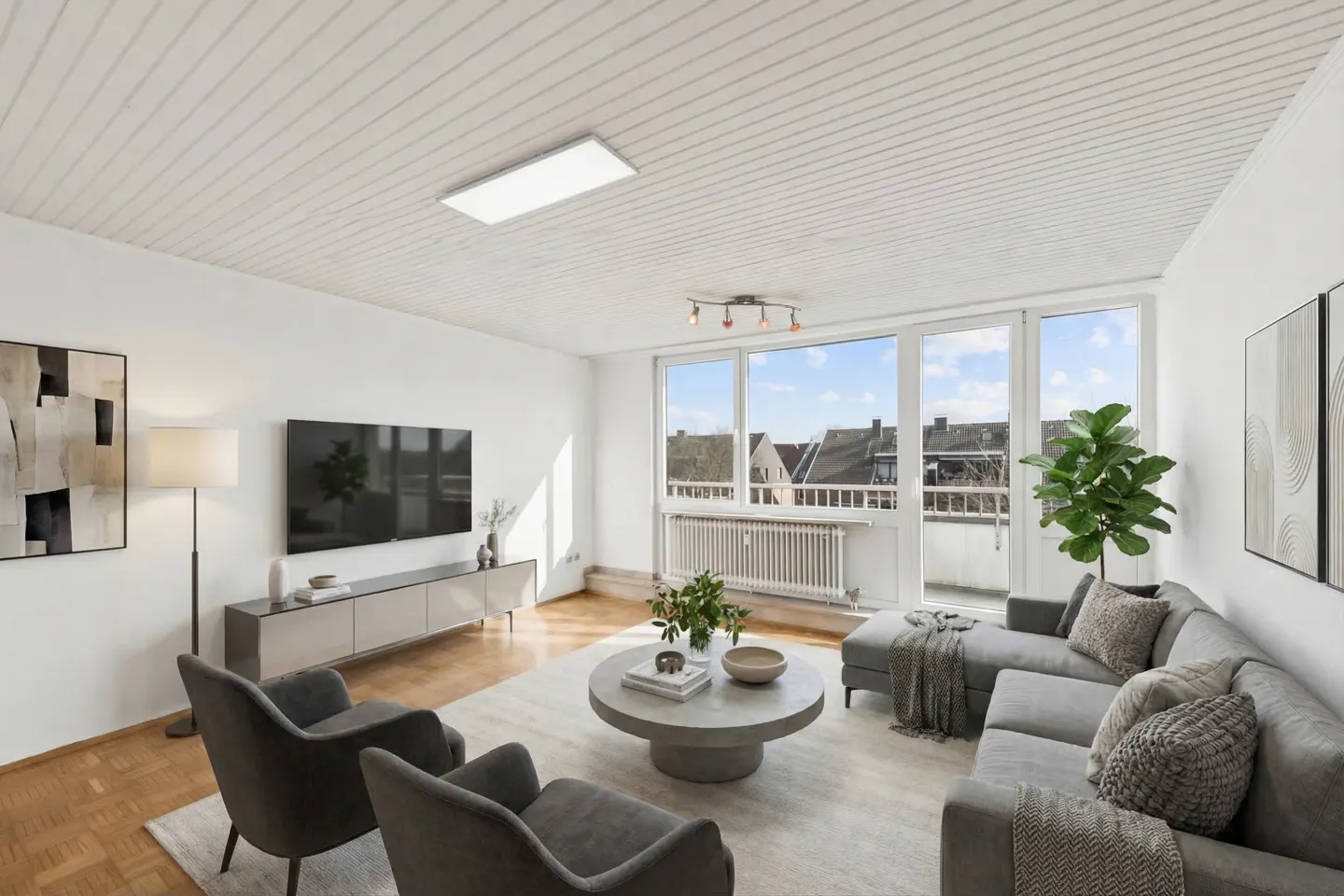 Bright living room with gray sofa, chairs, and a round coffee table on a light rug. Large windows offer a view of rooftops.