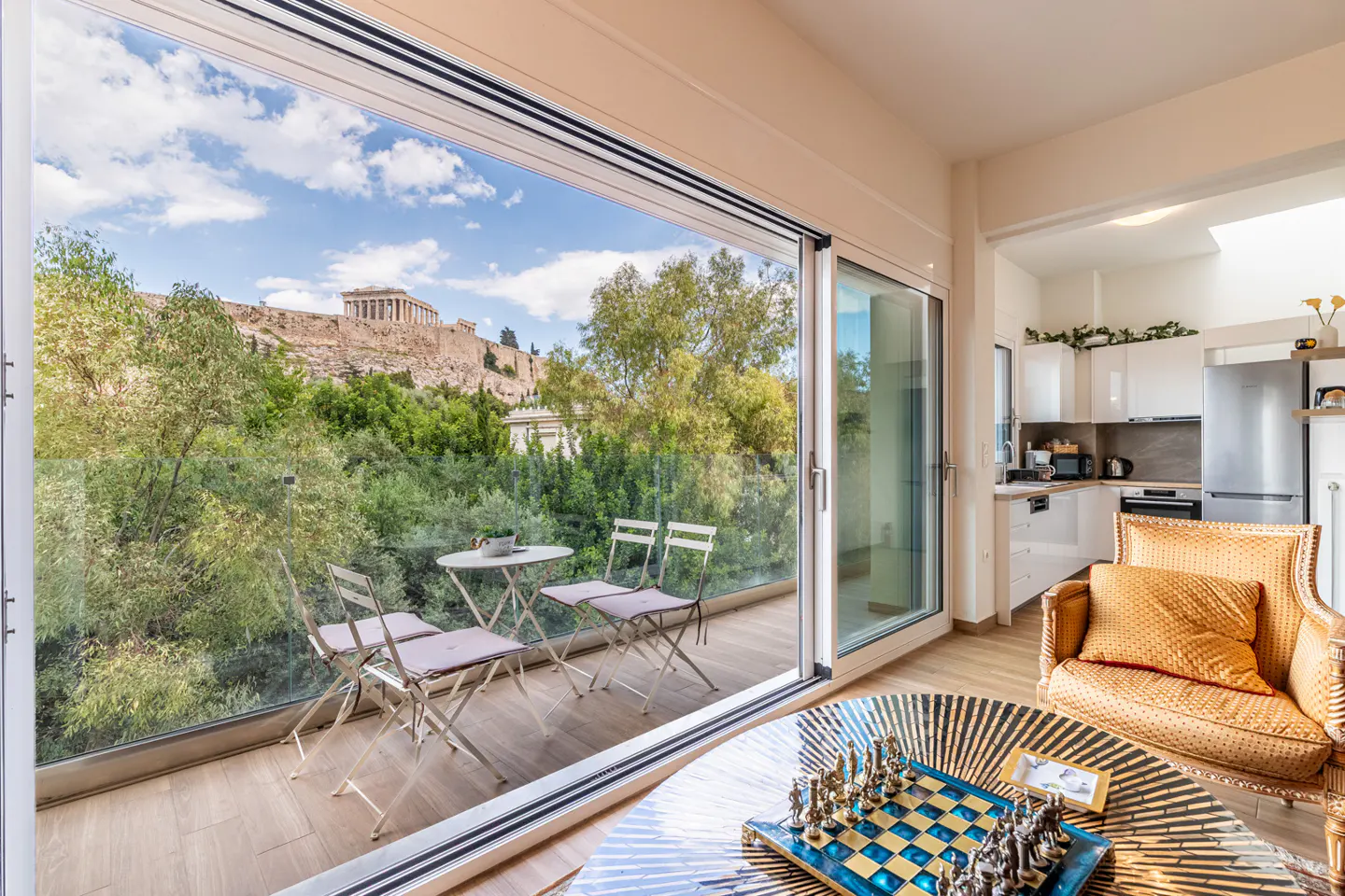 Living room with a chess set, gold armchair, and balcony overlooking the Parthenon in Athens through large glass doors.