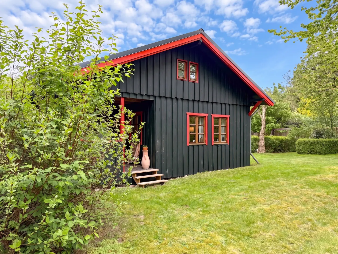 A black wooden house with red trim, two windows, and a small porch with steps sits on a green lawn.