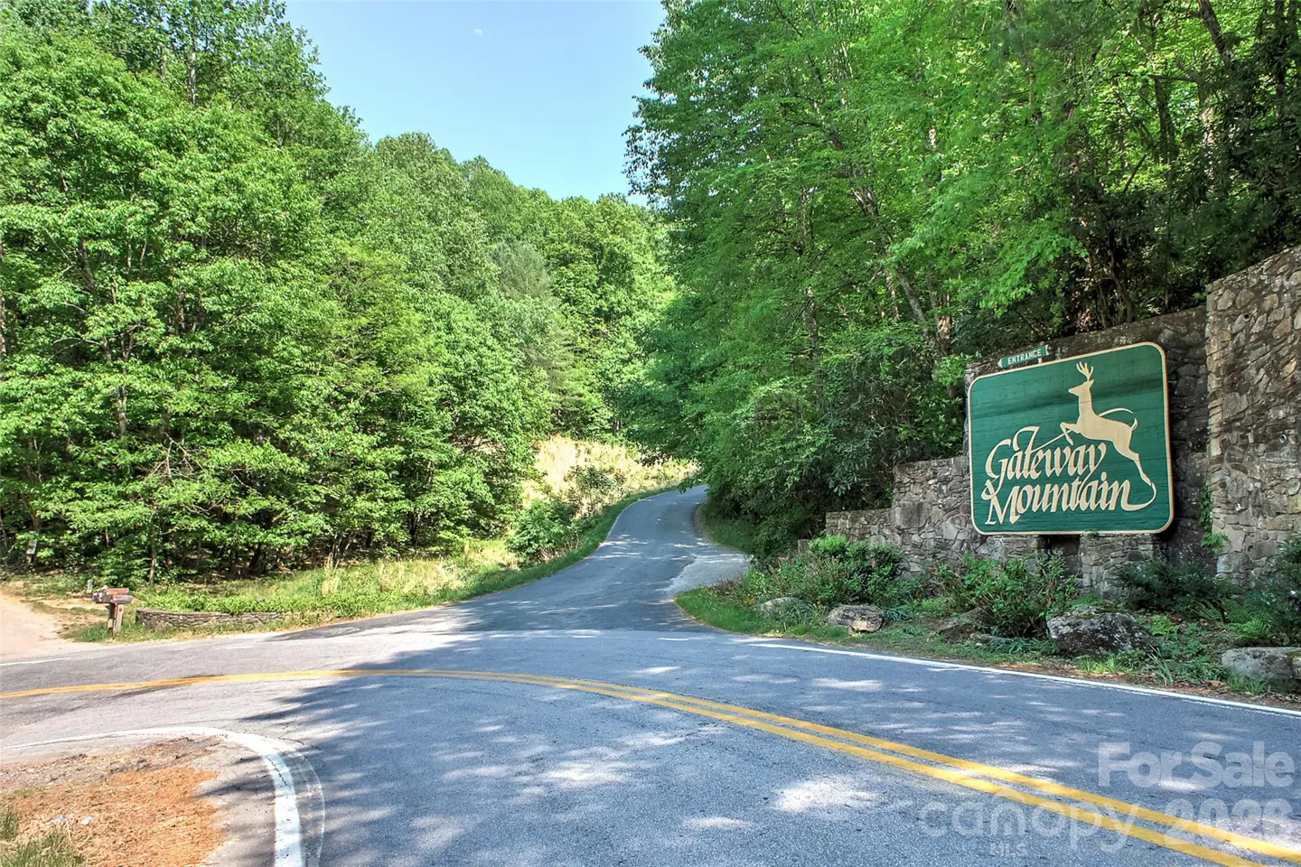 Road leading into Gateway Mountain, a green sign with a deer logo is on a stone wall. Trees line the road.
