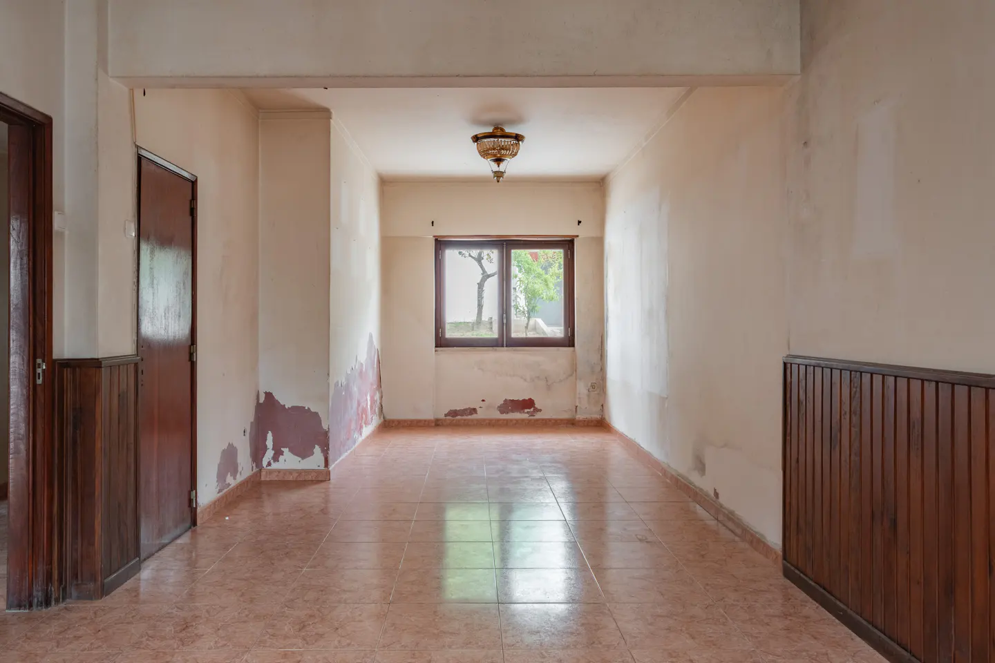 Empty room with peeling paint, brown tile floor, and wood paneling. A window shows a tree outside.