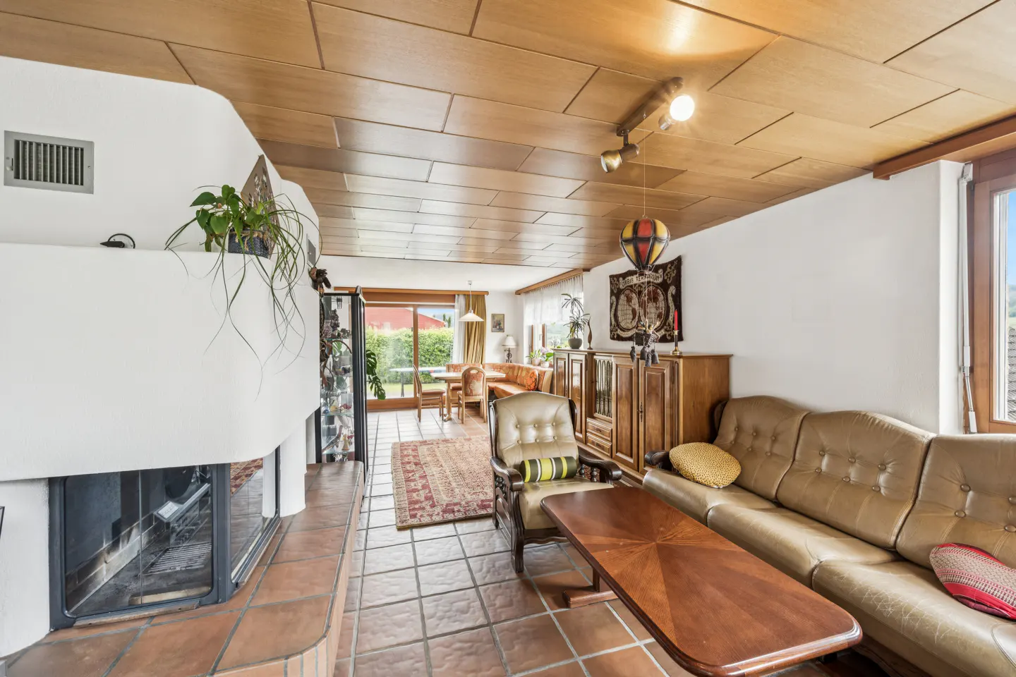 Living room with tiled floor, wood-paneled ceiling, fireplace, leather sofa, armchair, and dining area in the background.