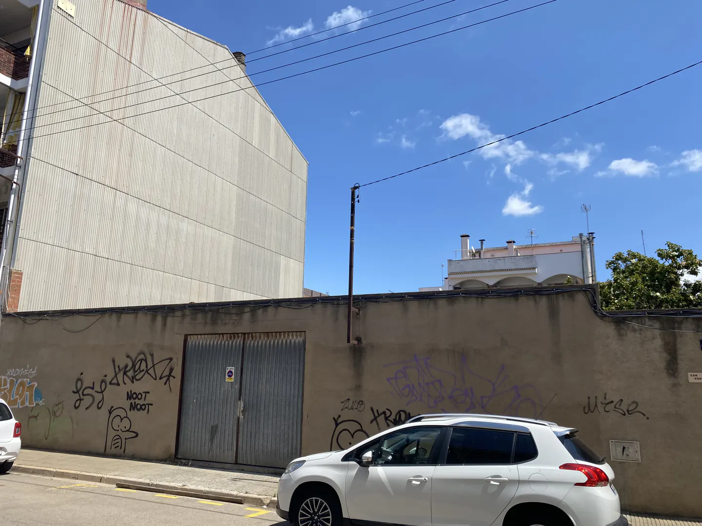 A white car parked in front of a beige wall with graffiti and a metal gate, under a clear blue sky.