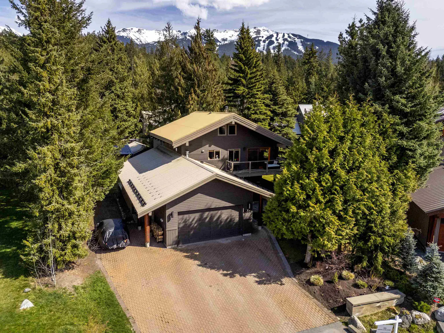 Aerial view of a brown two-story house with a garage, surrounded by green trees and snow-capped mountains in the background.