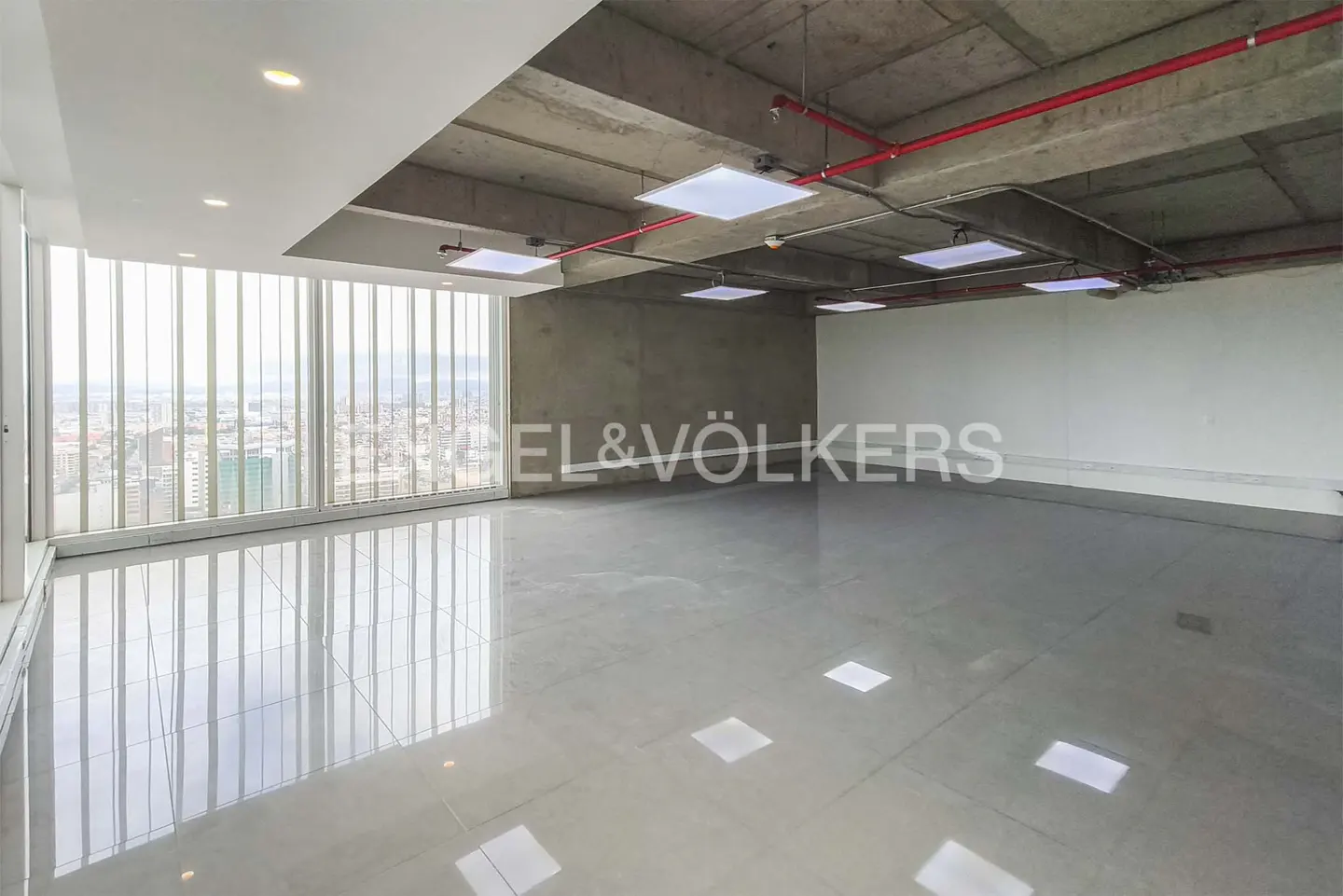 Empty office space with gray tile floor, concrete ceiling with red pipes, and a large window overlooking a city.