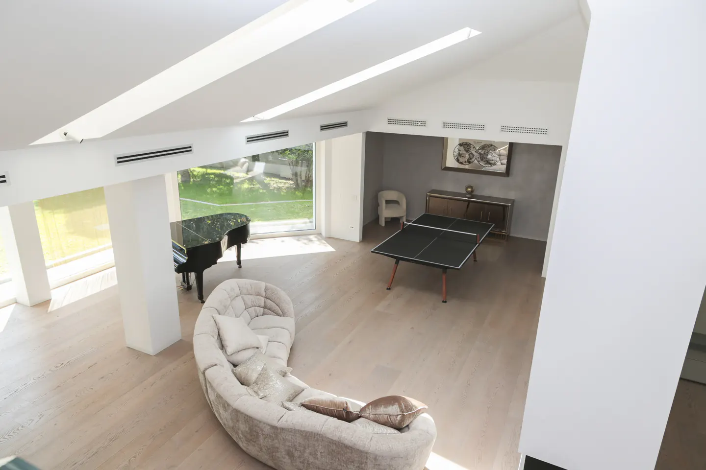 Bright, airy room with a piano, ping pong table, and curved sofa. Skylights illuminate the light wood floor.