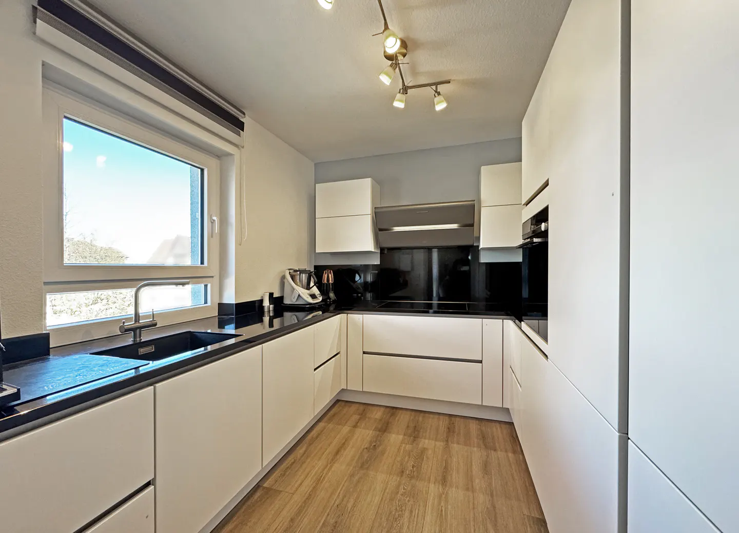 A modern kitchen with white cabinets, black countertops, and wood-look flooring. A window provides natural light.
