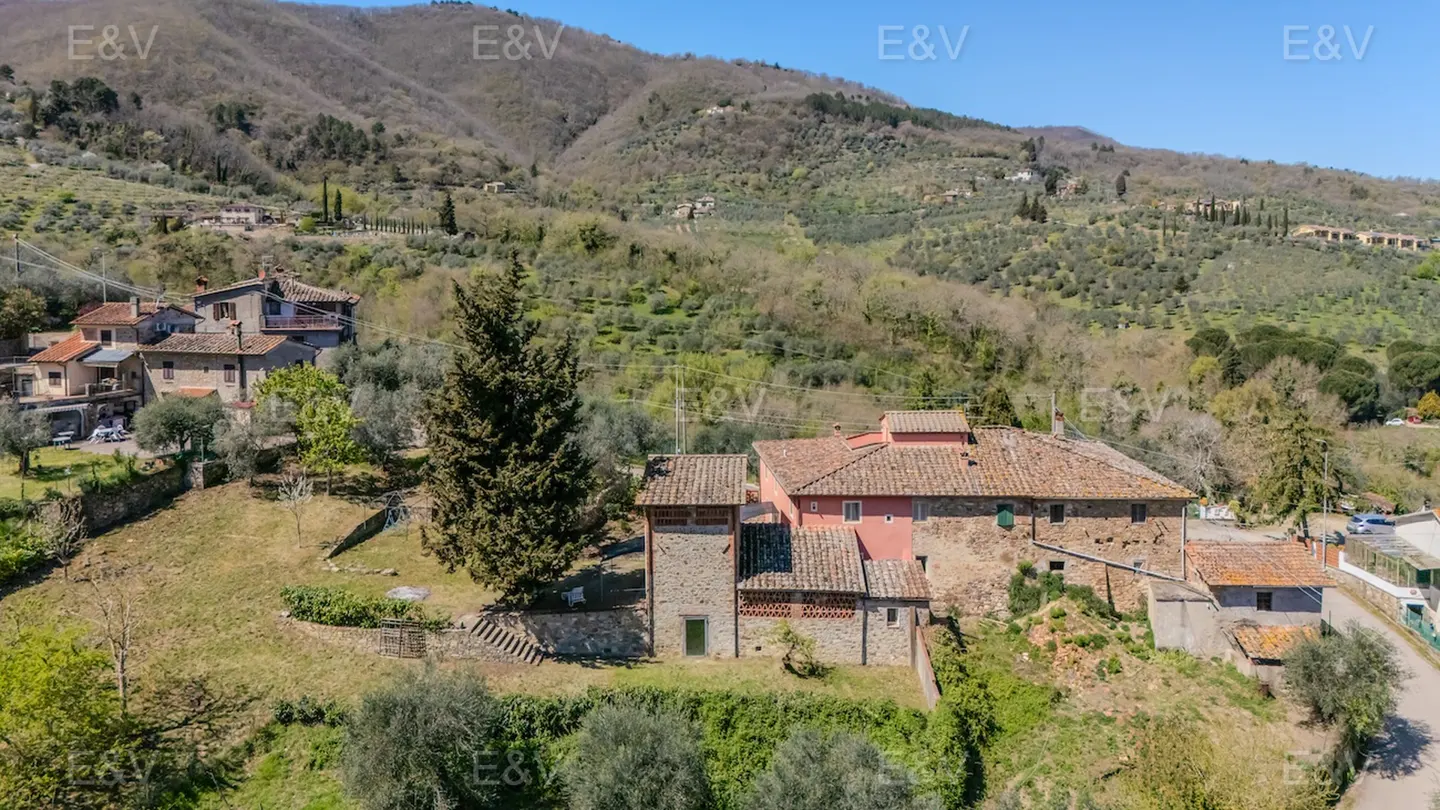 Aerial view of a pink house with a stone tower, surrounded by green hills and trees under a blue sky.