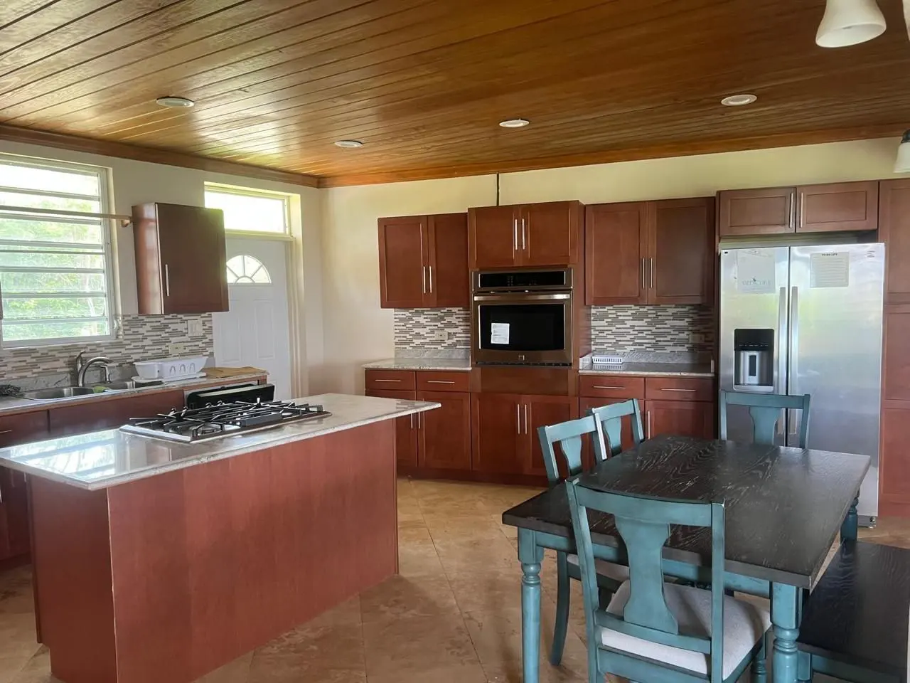 A kitchen with brown cabinets, stainless steel appliances, and a wooden ceiling. A blue table and chairs are in the foreground.
