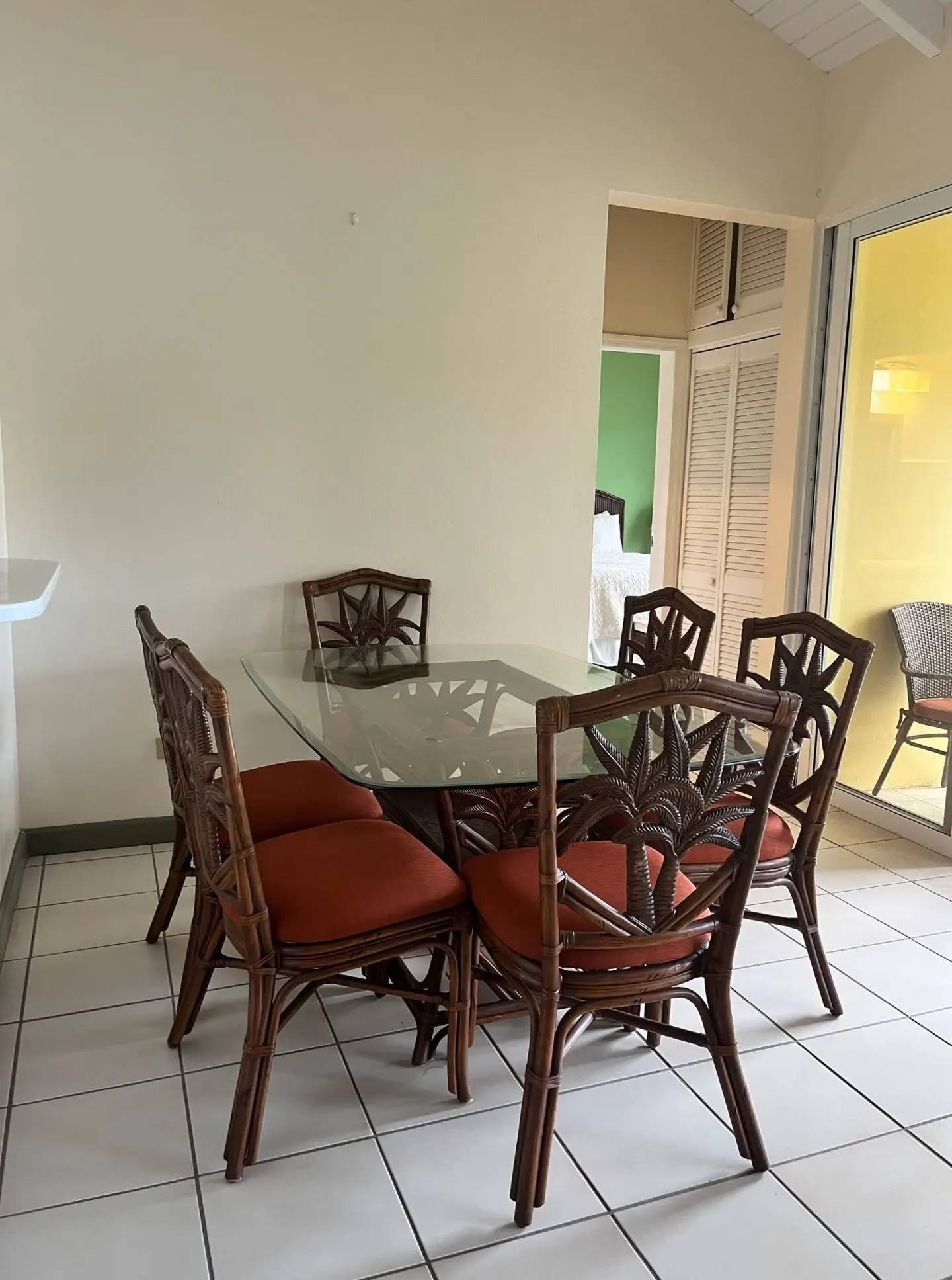 Dining area with a glass table and six brown wicker chairs with orange cushions. A bedroom and patio are visible in the background.