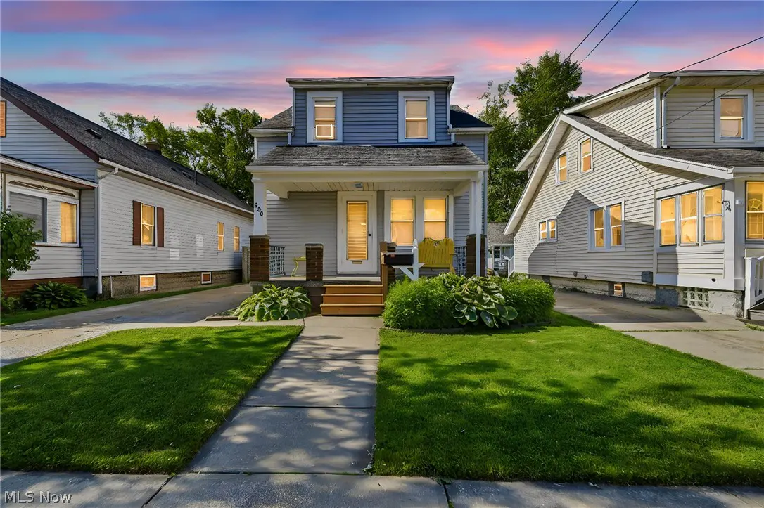 A blue two-story house with a front porch and yellow chairs, set between two homes under a vibrant sunset sky.