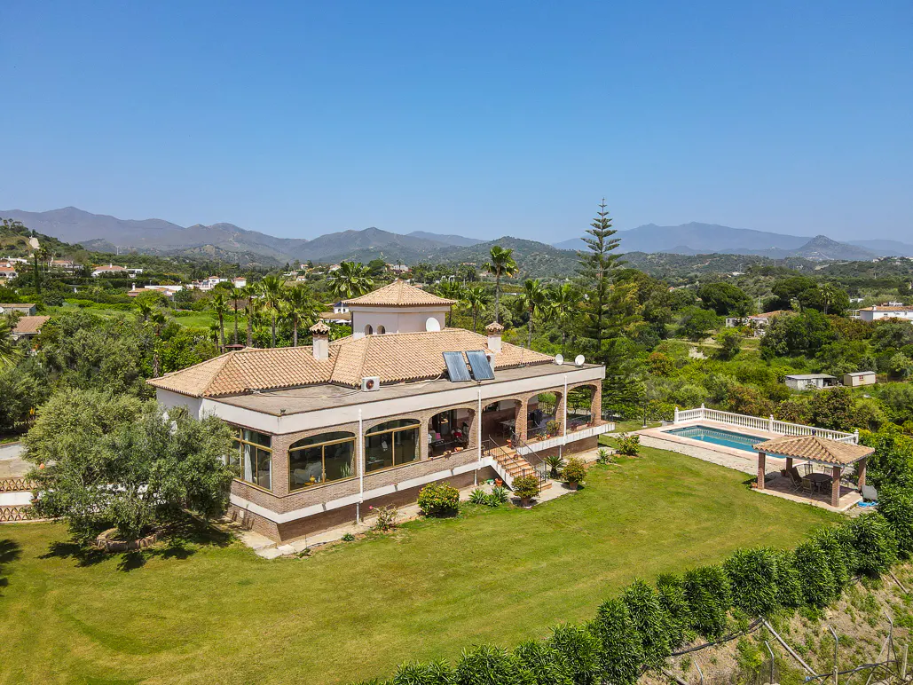 Aerial view of a large, tan house with a red tile roof, pool, and gazebo on a green lawn with trees and mountains in the background.