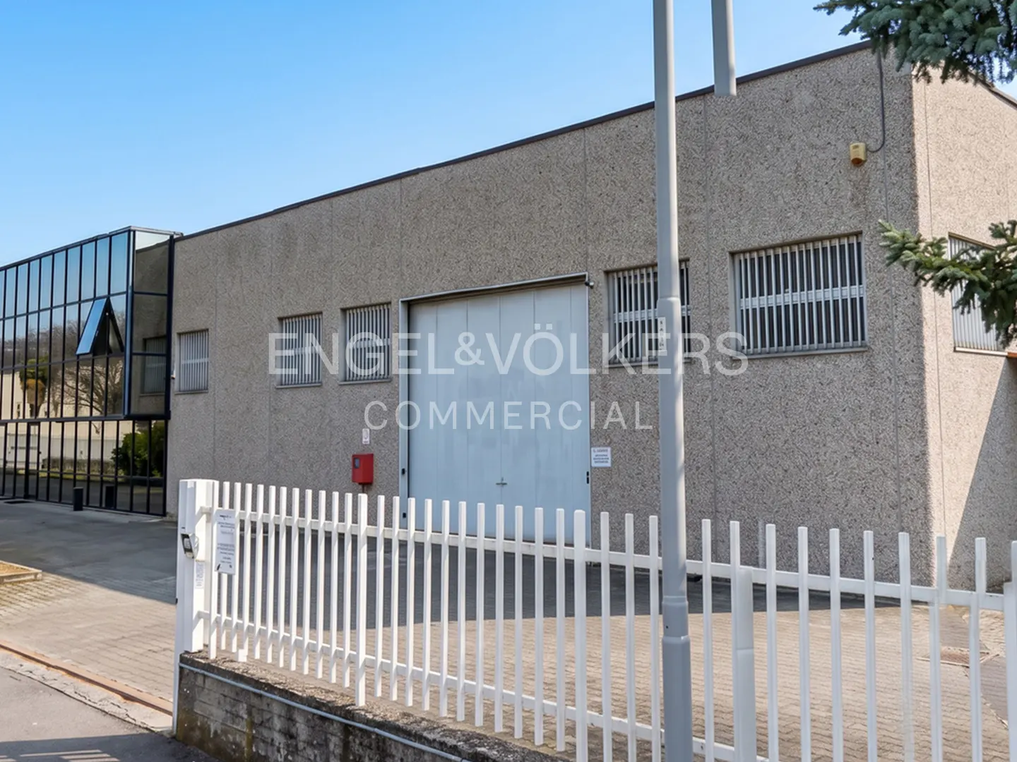 Commercial building with a large white door, barred windows, and a glass-walled section, behind a white picket fence.