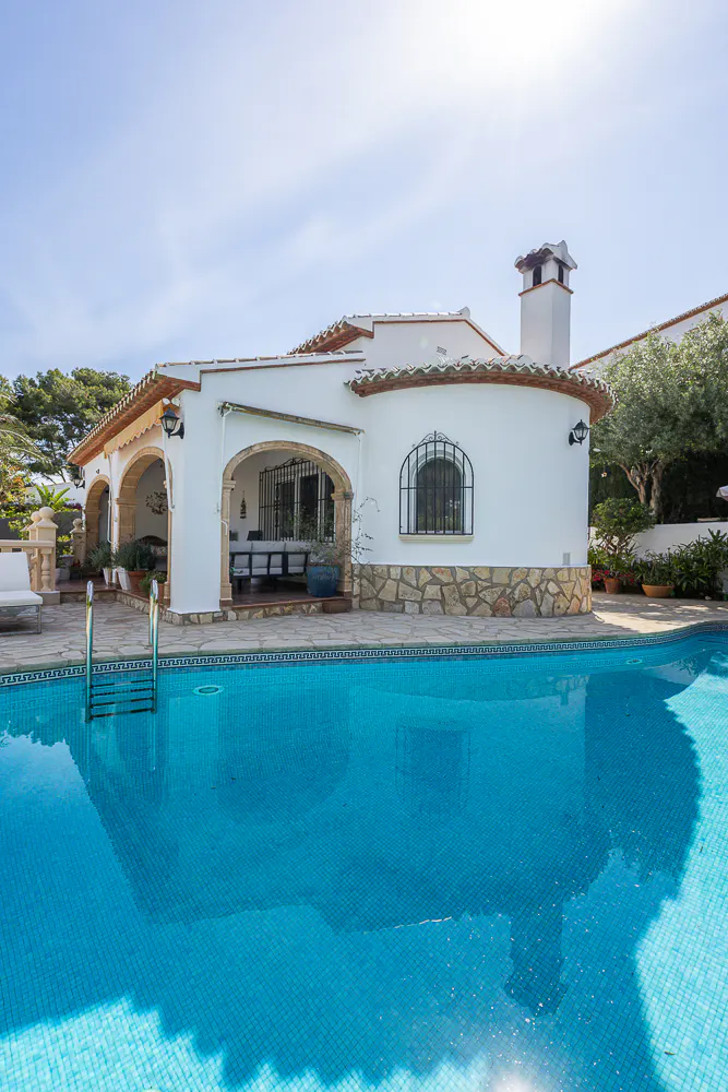 A white house with a red tile roof sits beside a blue tiled swimming pool on a sunny day.