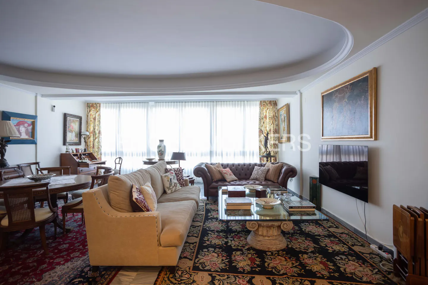 Living room with beige sofa, brown leather sofa, glass coffee table, and floral rug. Art on the walls and large windows.
