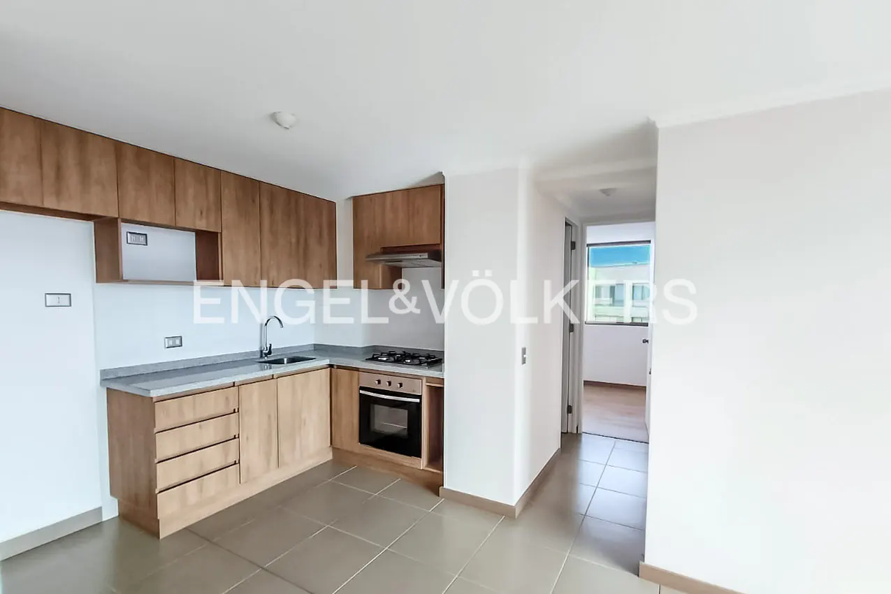 Modern kitchen with light wood cabinets, grey countertops, built-in oven, and tiled floors, leading to a hallway.