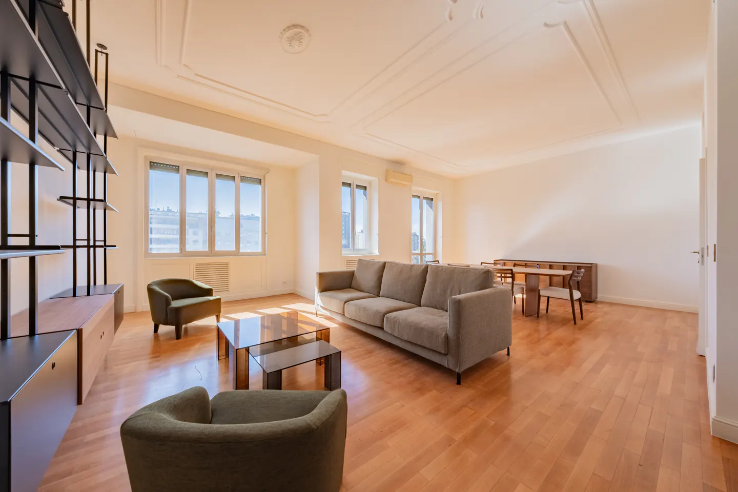 Bright living room with wood floors, white walls, and large windows. A gray sofa, green chairs, and glass tables furnish the space. A dining table sits in the background.