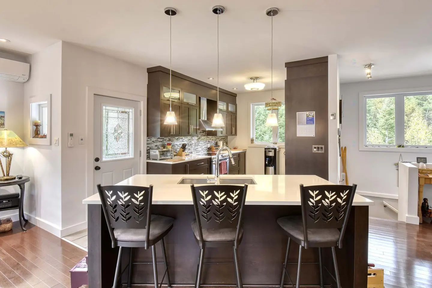 Open concept kitchen with dark cabinets, white countertops, and three black bar stools with leaf cutouts. Hardwood floors and pendant lighting.