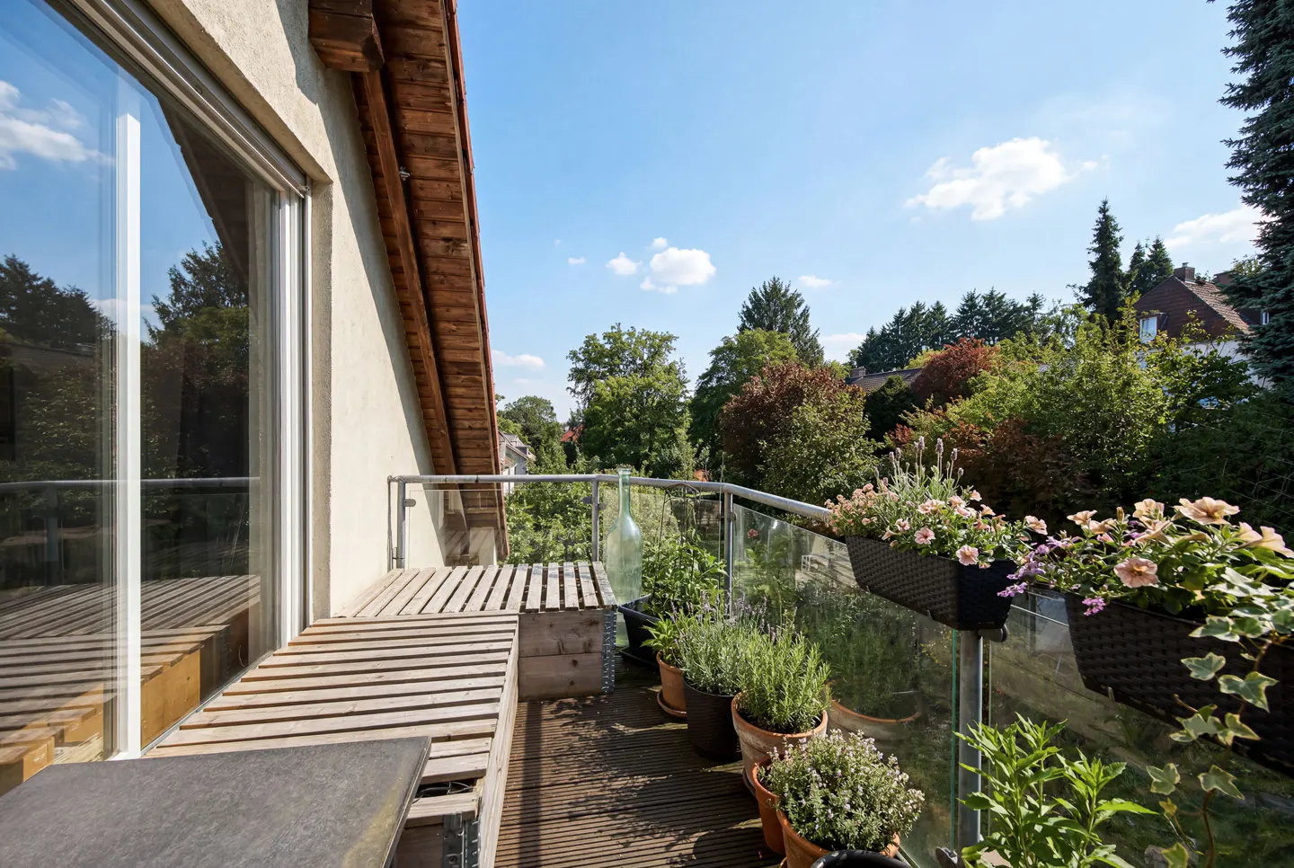 A sunny balcony with wood floors, a bench, and potted plants. A glass railing overlooks lush green trees.