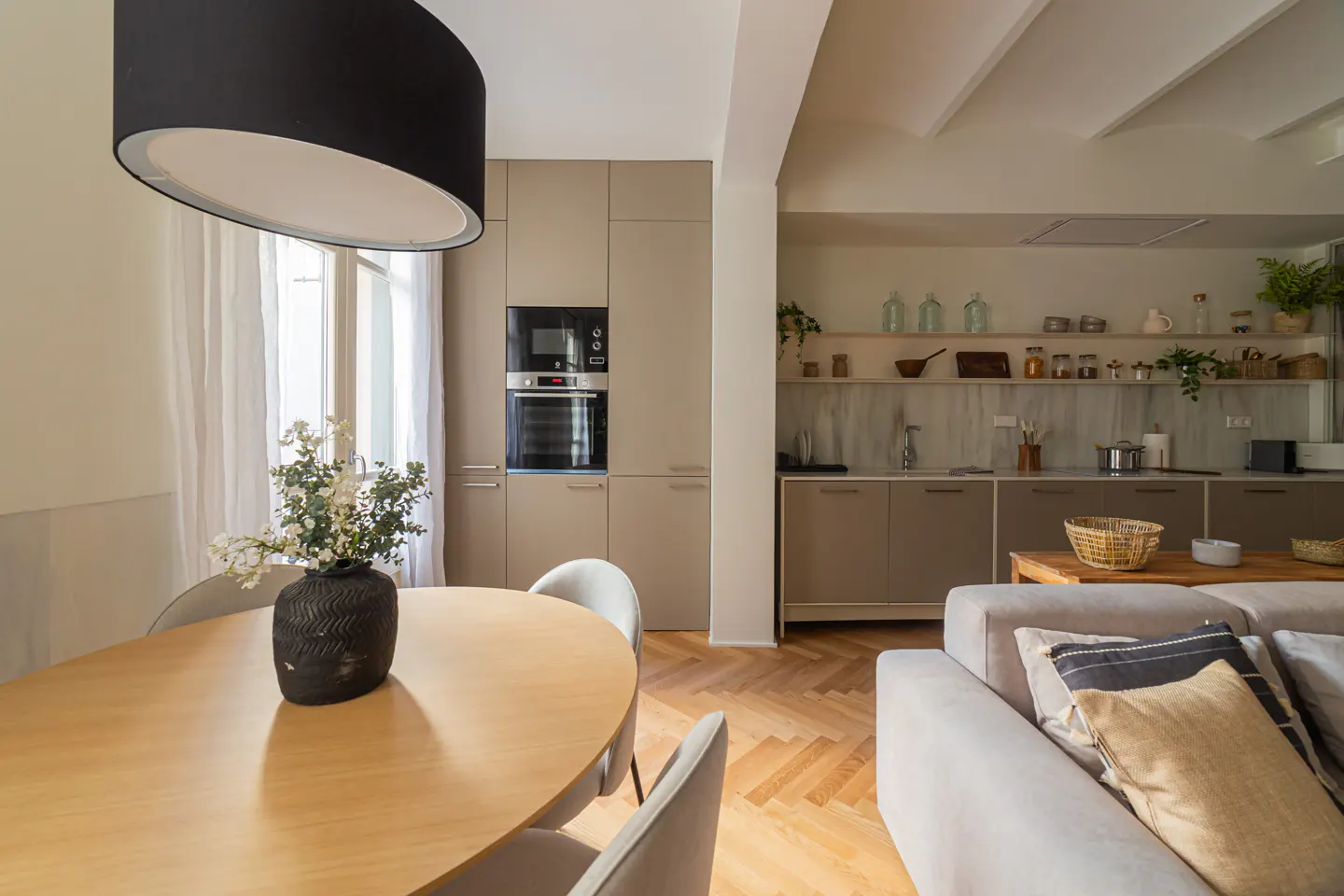 Open-concept kitchen and living room with wood floors, beige cabinets, and a round wood table with a black vase of white flowers.