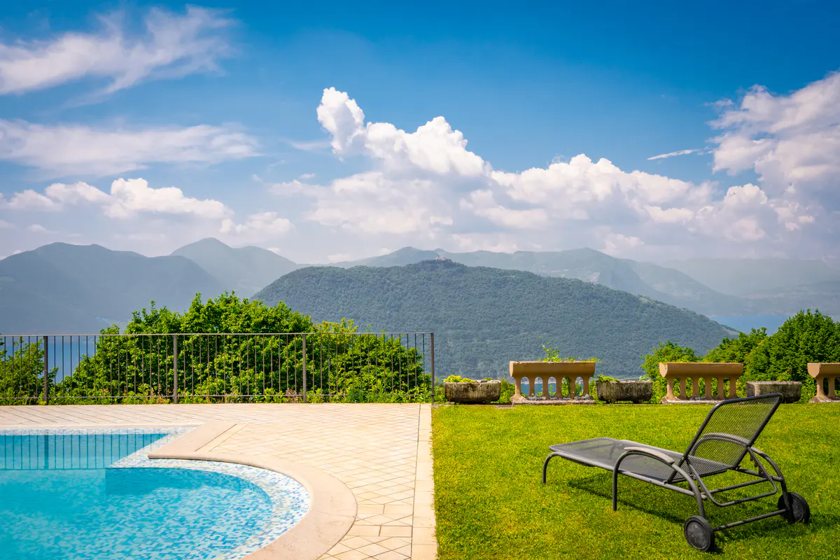 Outdoor pool with lounge chair on green grass, stone planters, and mountain views under a blue sky with white clouds.