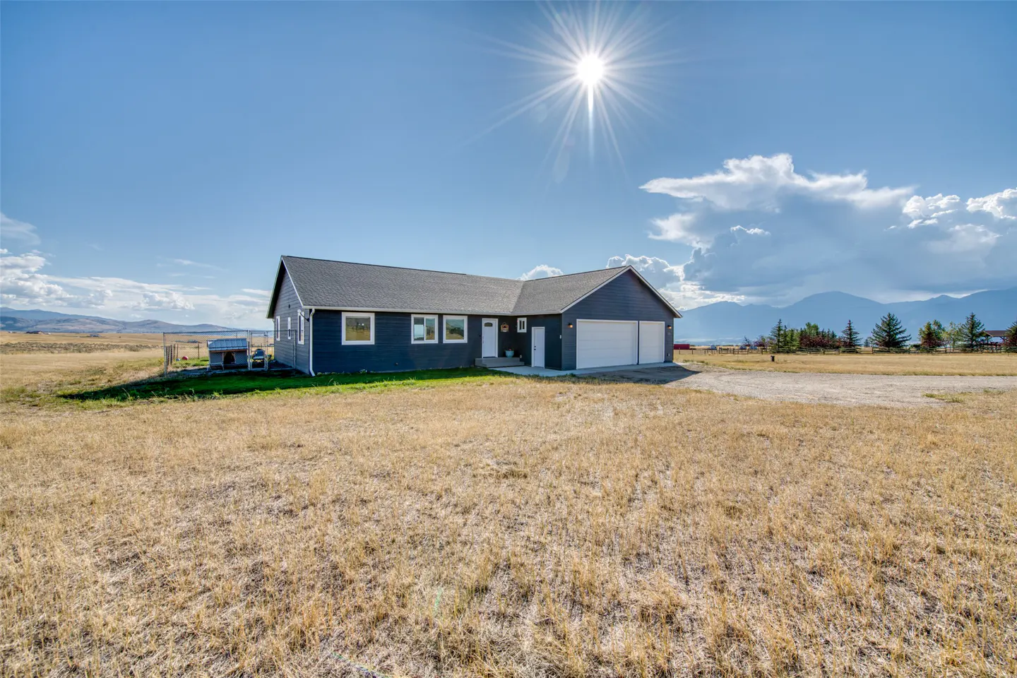Dark gray ranch-style home with a two-car garage set in a vast, dry grassy field under a bright sun and mountain backdrop.