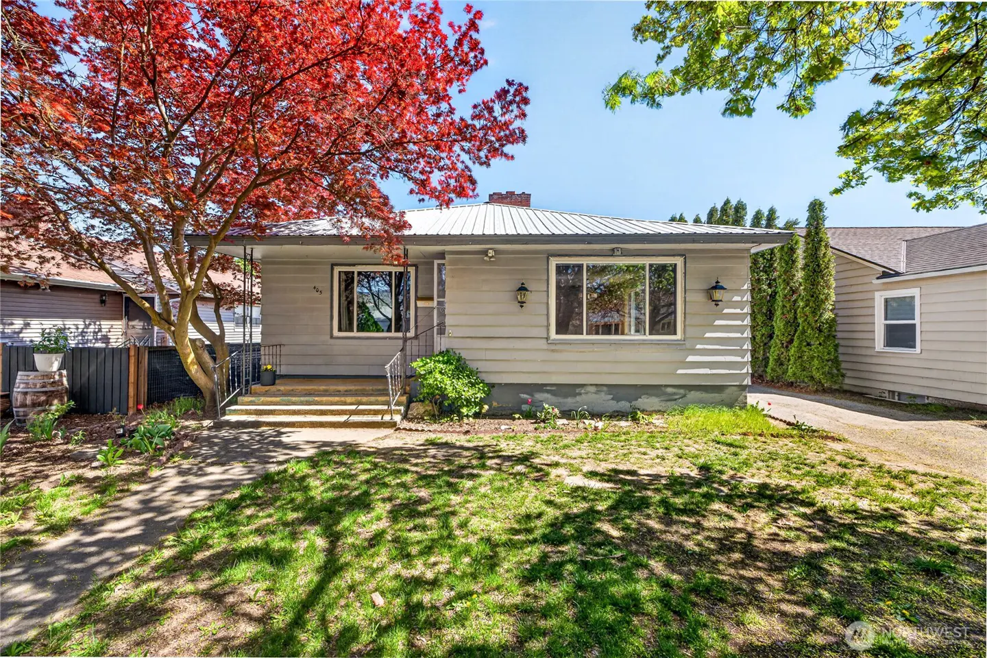 A gray, one-story house with a metal roof, small porch, and a large red-leafed tree in the front yard.