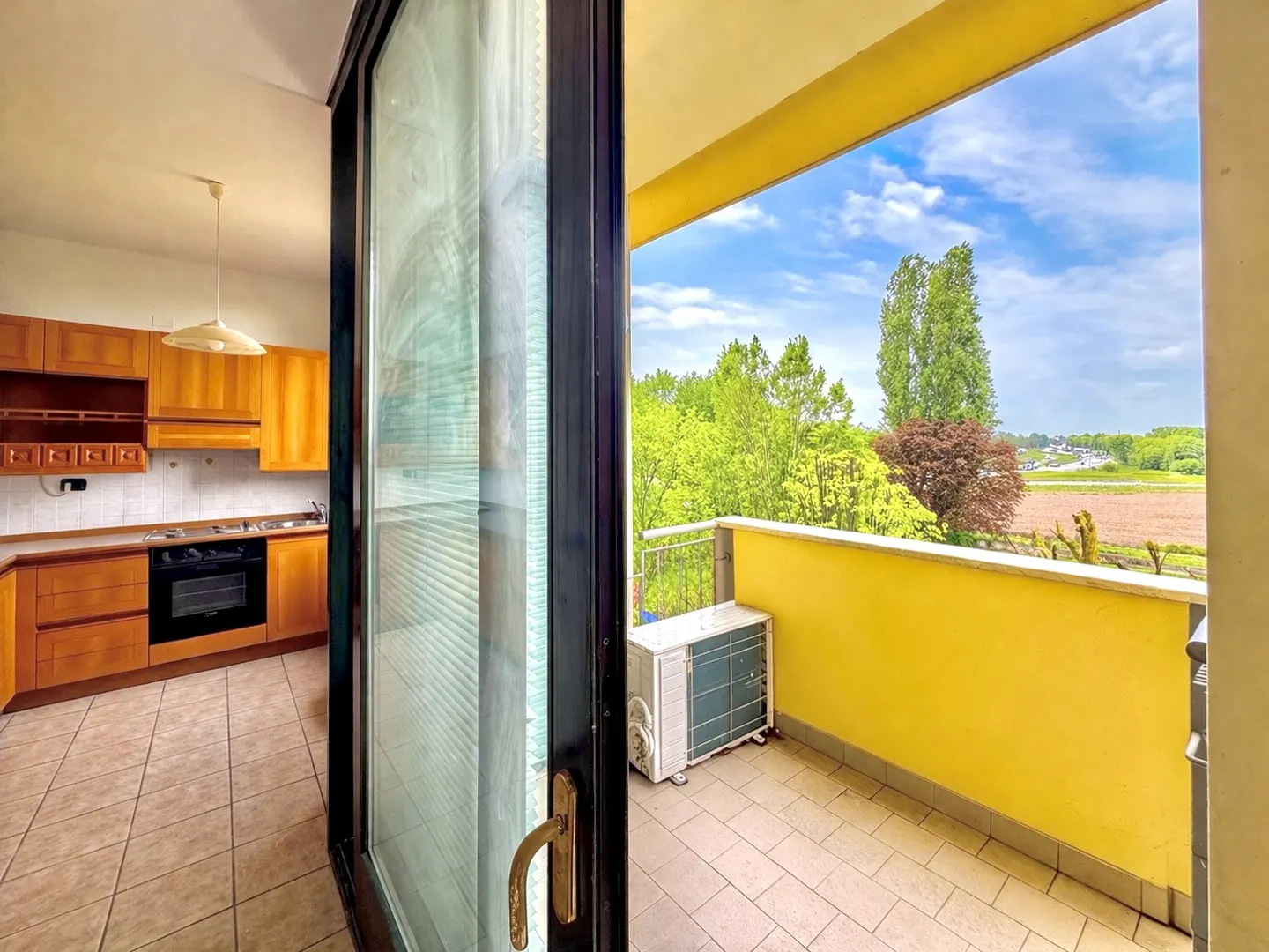 View from an open sliding glass door to a kitchen and a yellow balcony with an AC unit and a view of trees and a field.