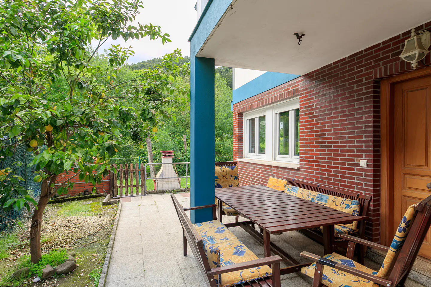 Outdoor patio with a wooden table, benches, and chairs. A brick house with a blue pillar and a tree with yellow fruit are visible.