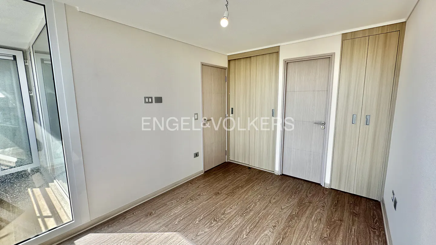 A bright, empty bedroom with wood floors, white walls, and built-in closets. A glass door leads to a balcony.