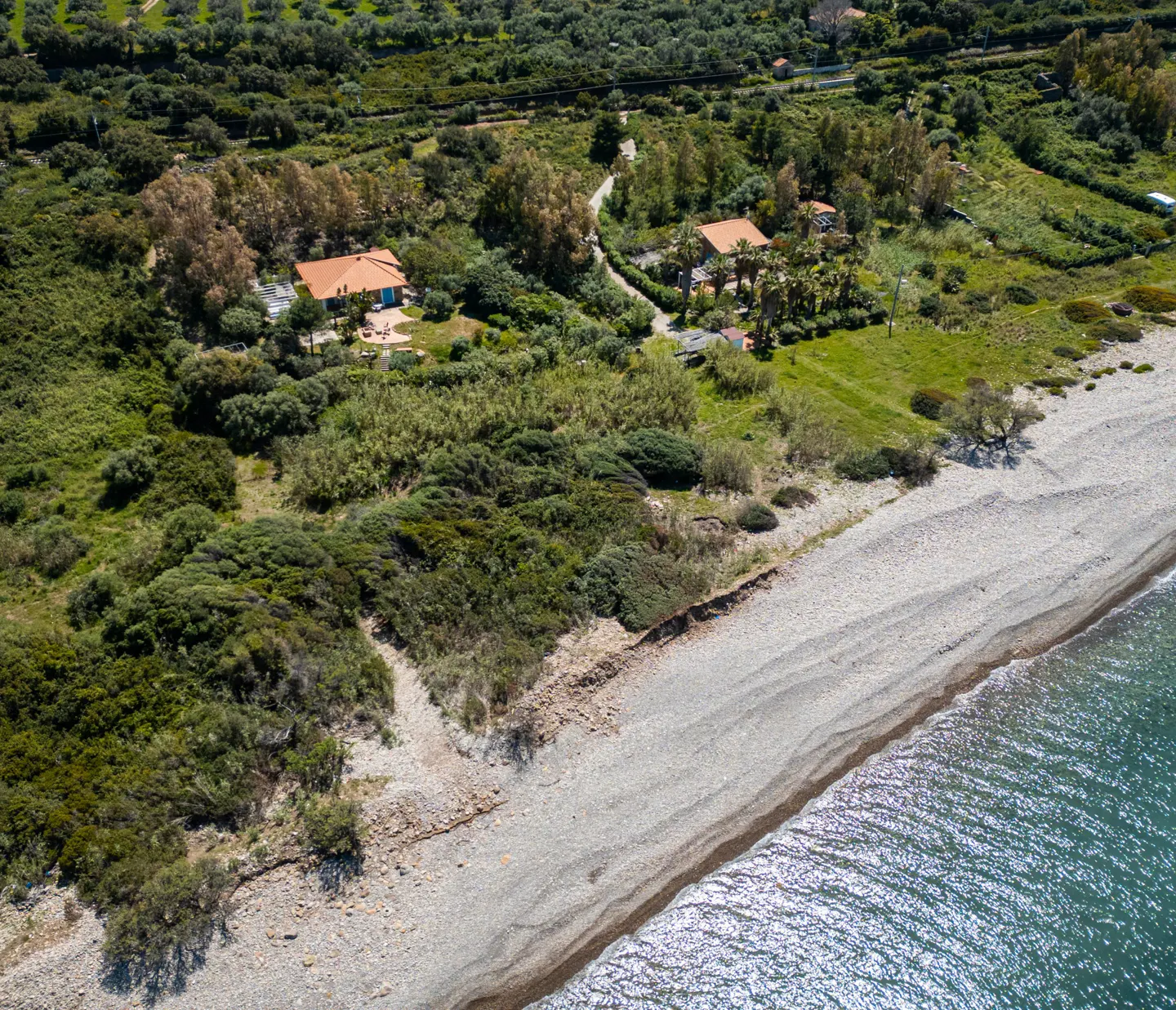 Aerial view of two houses with red tile roofs nestled in lush greenery near a pebble beach and blue ocean.