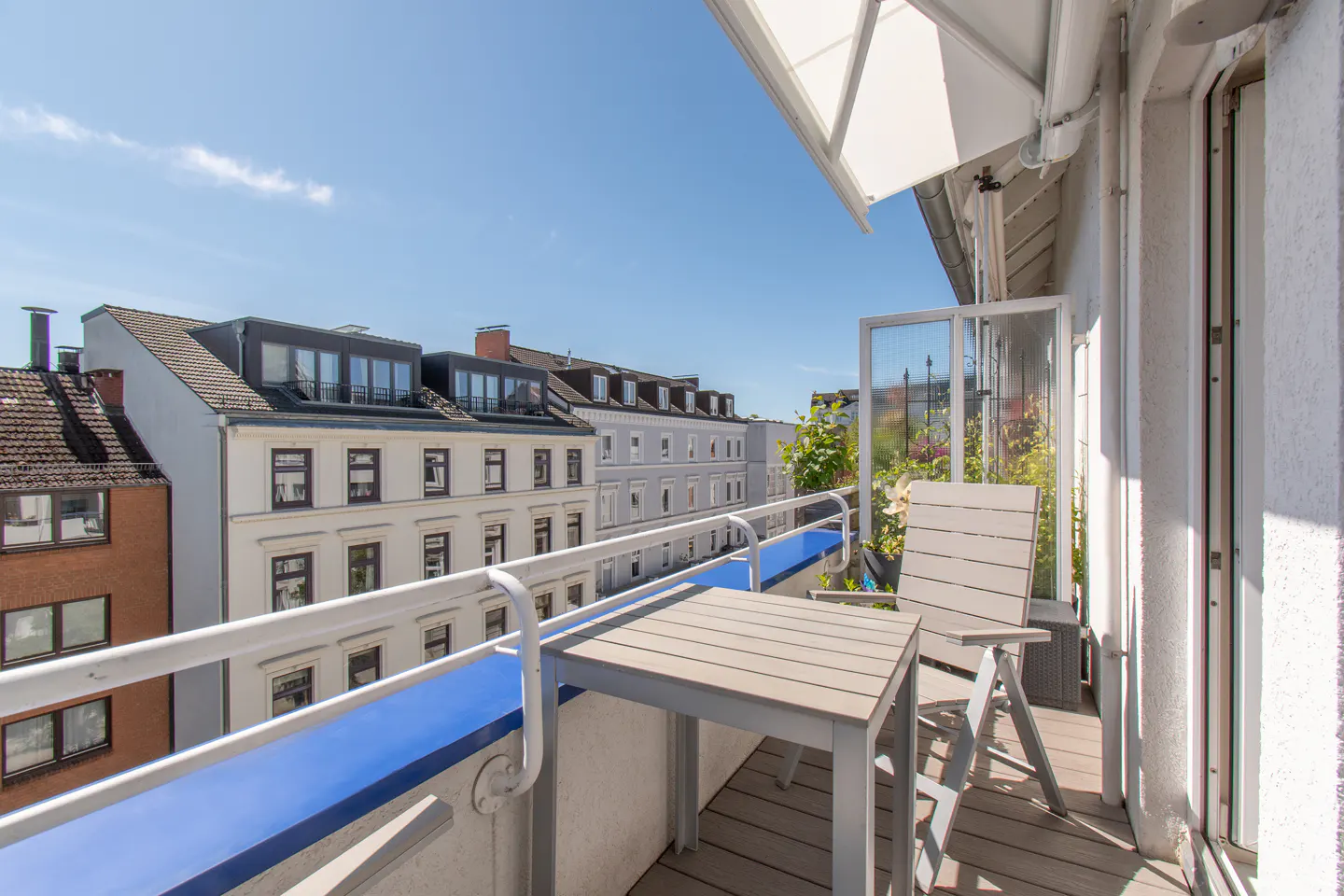 Sunny balcony with a small table and chair, overlooking a city street with multi-story apartment buildings.