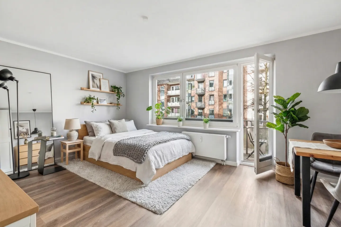 Bright studio apartment with a bed, dining table, and balcony. Gray walls, wood floors, and large windows create an airy feel.