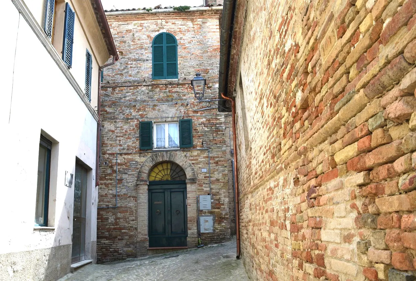 A narrow cobblestone street between brick buildings with green shutters and doors.