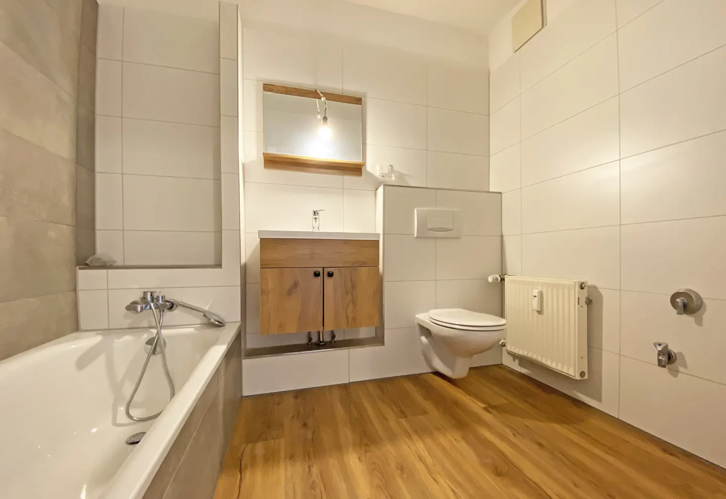 Bright bathroom with white tile walls, wood-look floors, and a white bathtub. A wooden vanity with a mirror hangs above a toilet and radiator.