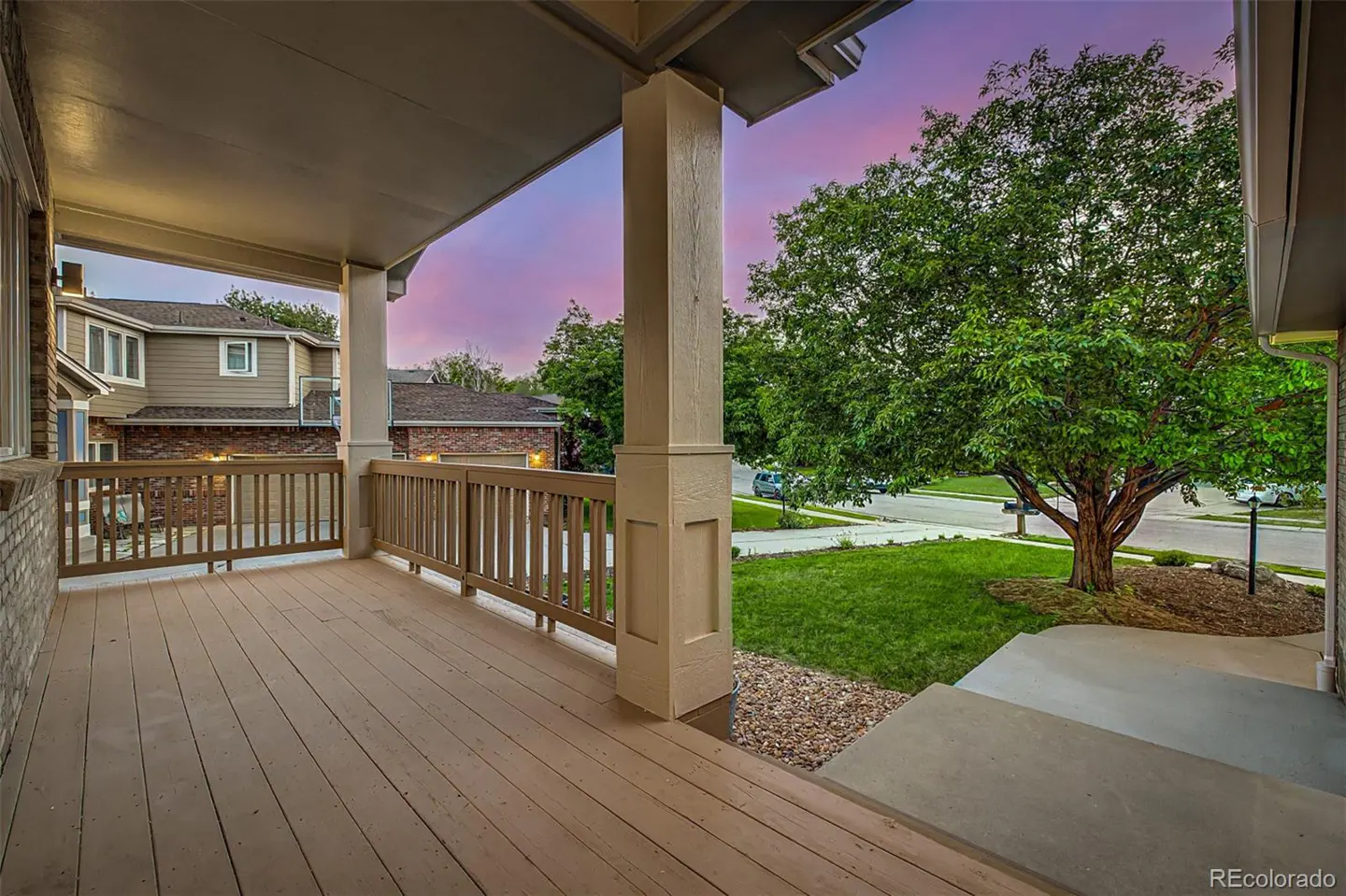 Covered front porch with wood floor and railing. A large tree and green lawn are visible. The sky is pink and purple.