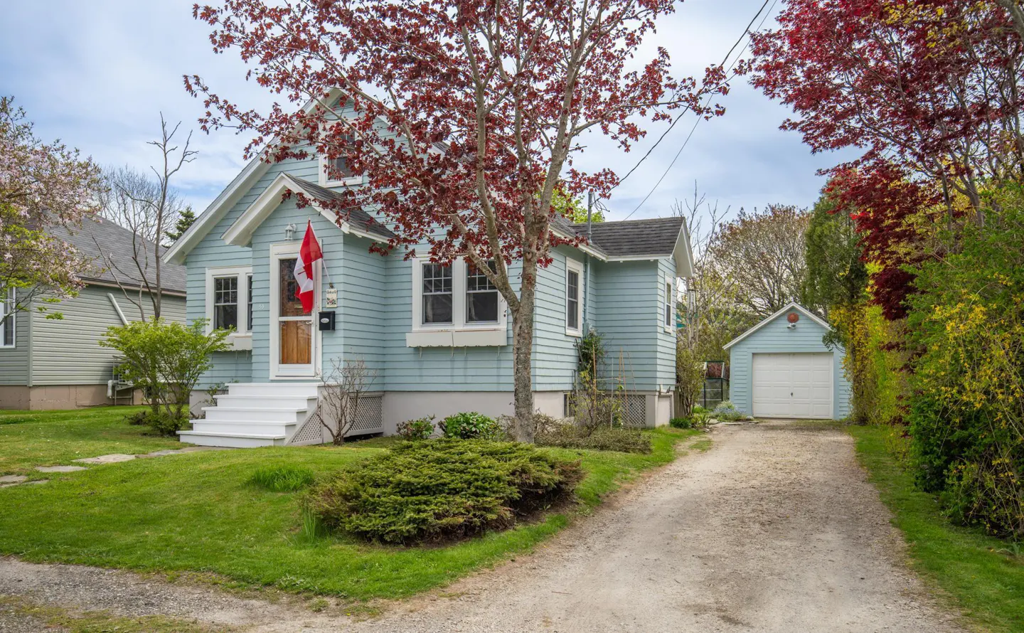 Light blue house with white trim, a Canadian flag, and a detached garage on a gravel driveway.