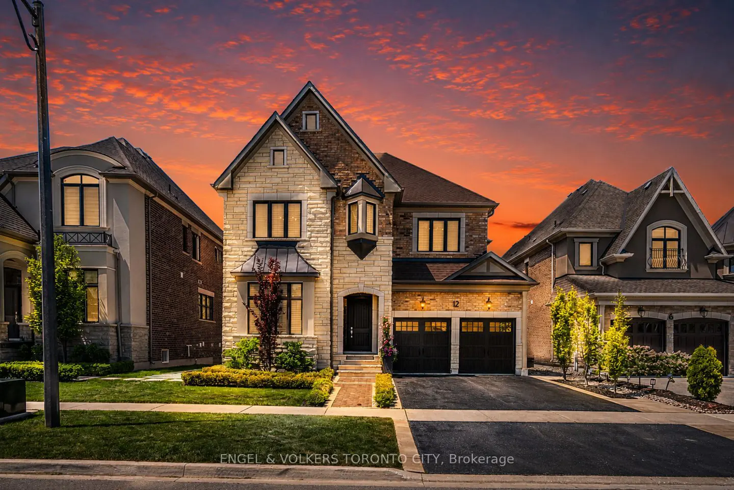 A two-story stone and brick suburban home at sunset with a vibrant orange sky, double garage, and manicured front lawn.