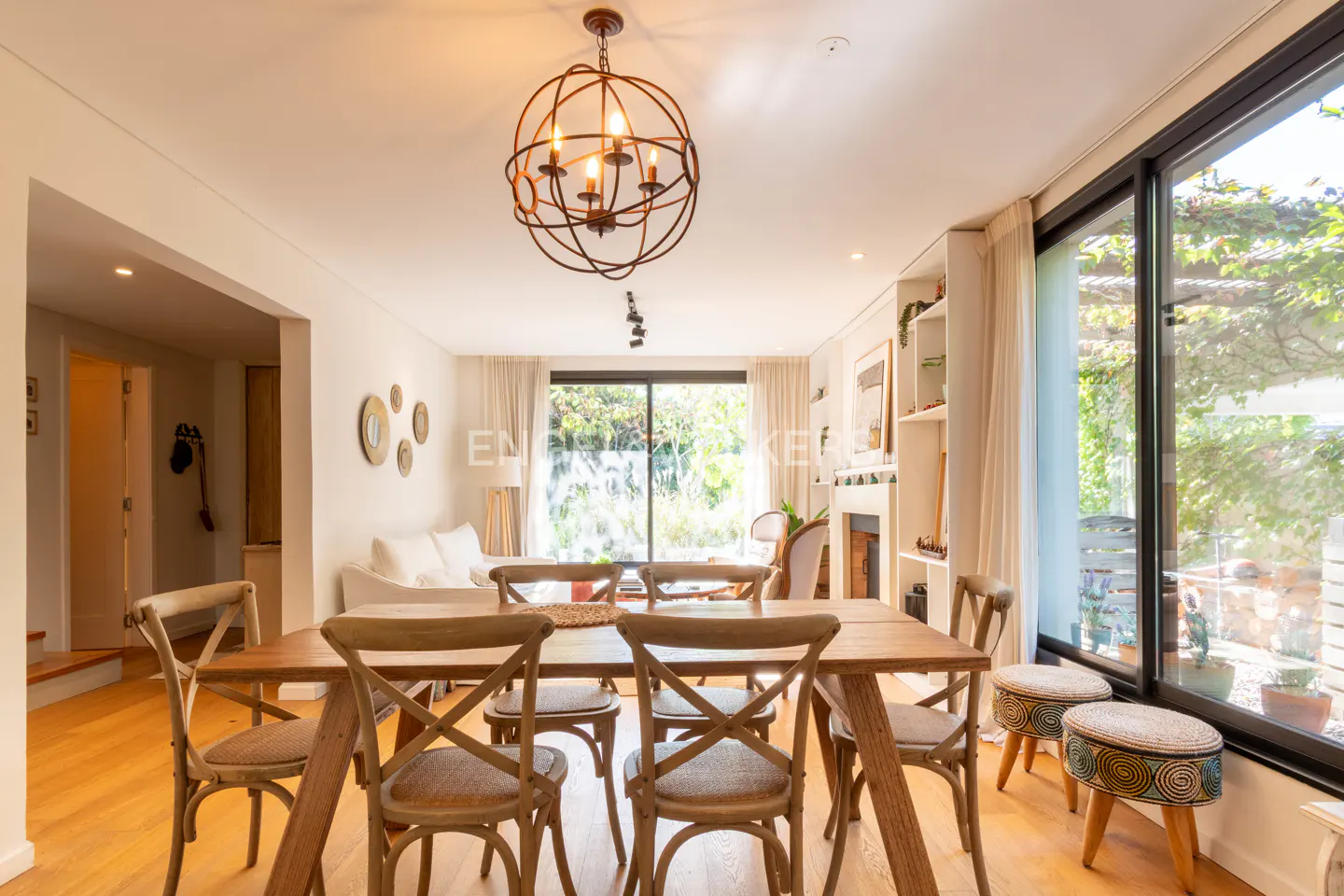A bright dining room with a wooden table and chairs under a spherical chandelier. Large windows overlook a lush garden.