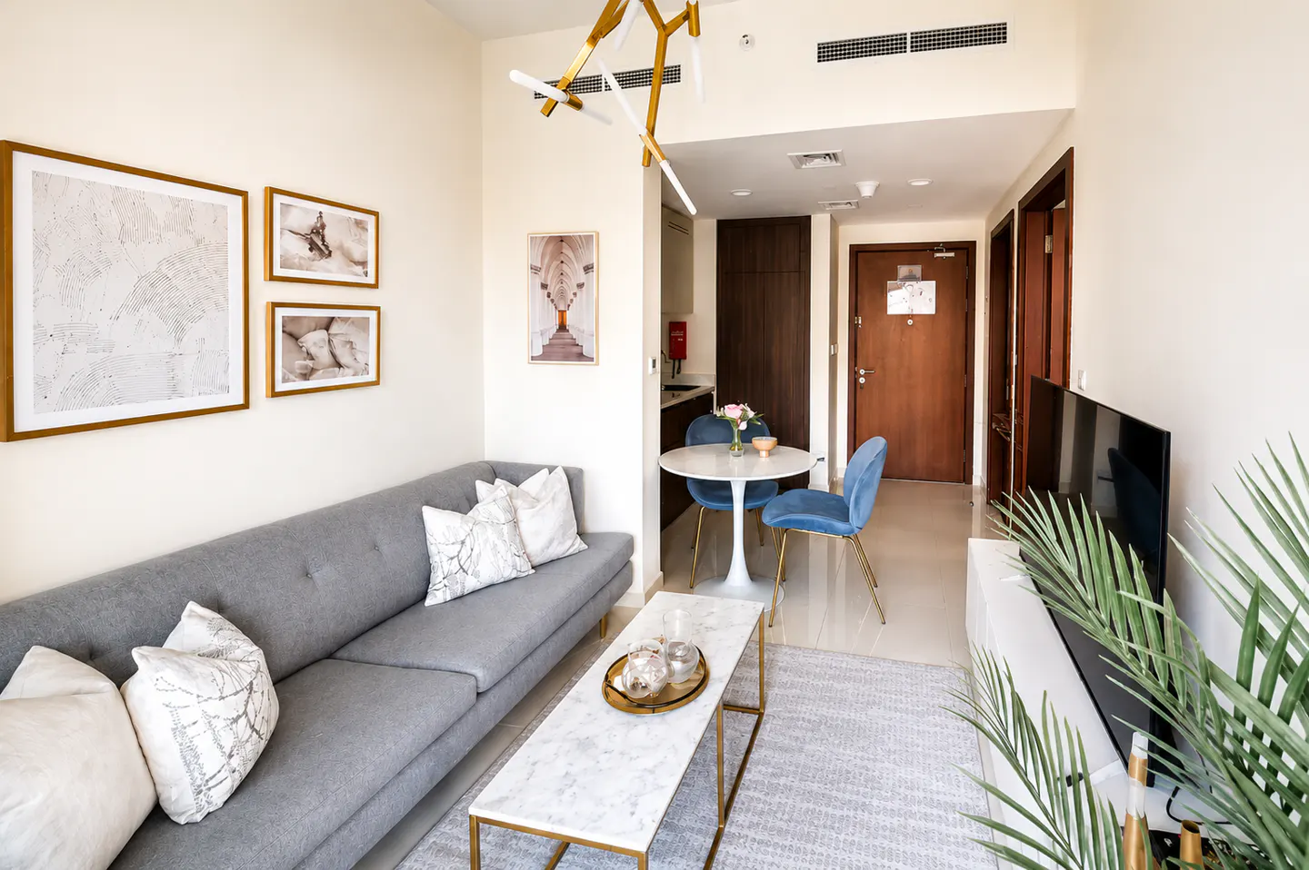 Living room with gray sofa, marble coffee table, and blue chairs around a white table. Artwork adorns the walls, and a modern chandelier hangs above.