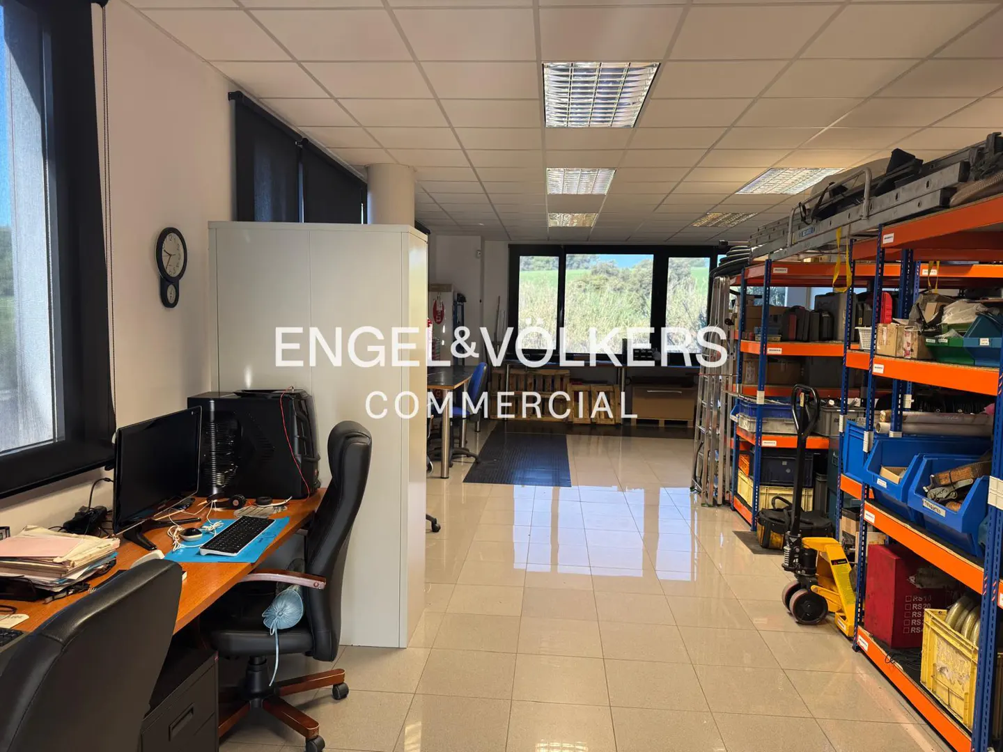 Bright office space with desks, computers, and shelving units filled with supplies. Large windows offer a view of greenery.