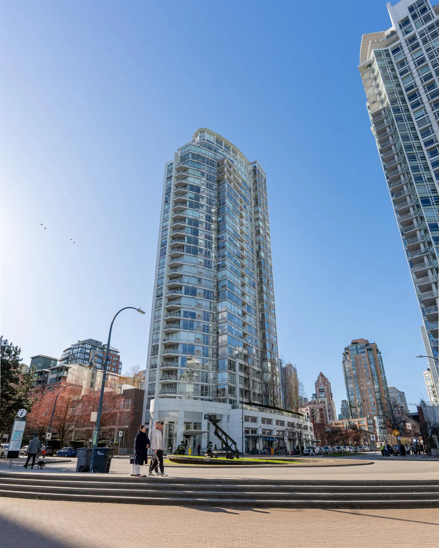 Exterior view of a tall, modern glass condo building on a sunny day with blue sky. People walk in the plaza.