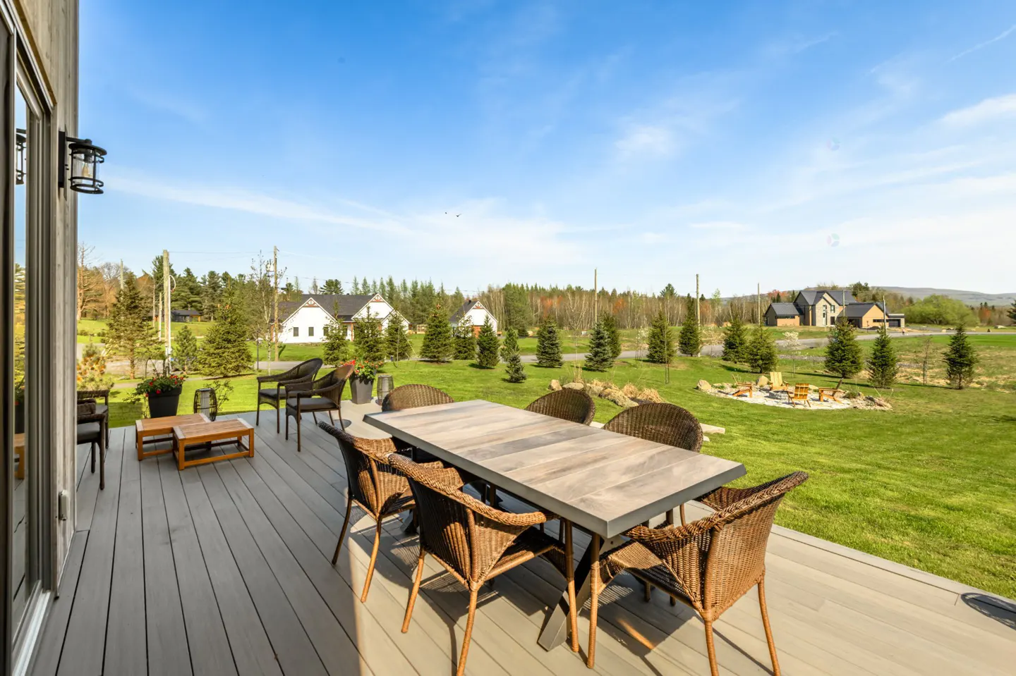 Outdoor patio with a wooden table and wicker chairs on a grey deck, overlooking a green lawn and houses under a blue sky.