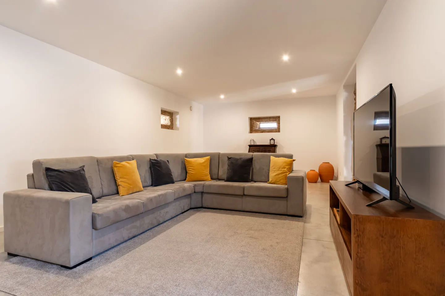 Living room with a gray sectional sofa, yellow and black pillows, a gray rug, and a TV on a wooden stand. White walls and recessed lighting.