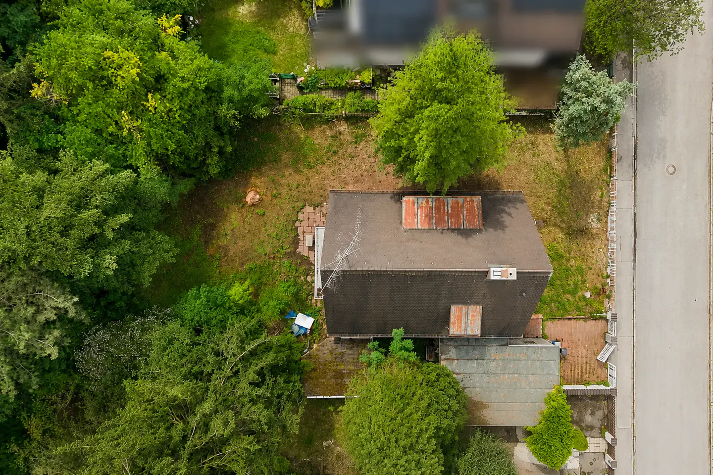 Aerial view of a house with a dark roof and rusty skylights, surrounded by lush green trees and a large grassy yard.
