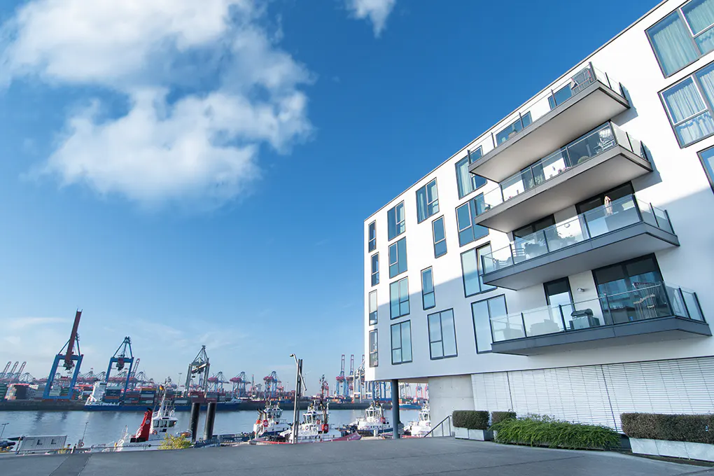 Modern white building with balconies overlooks a harbor with cranes and boats under a blue sky.