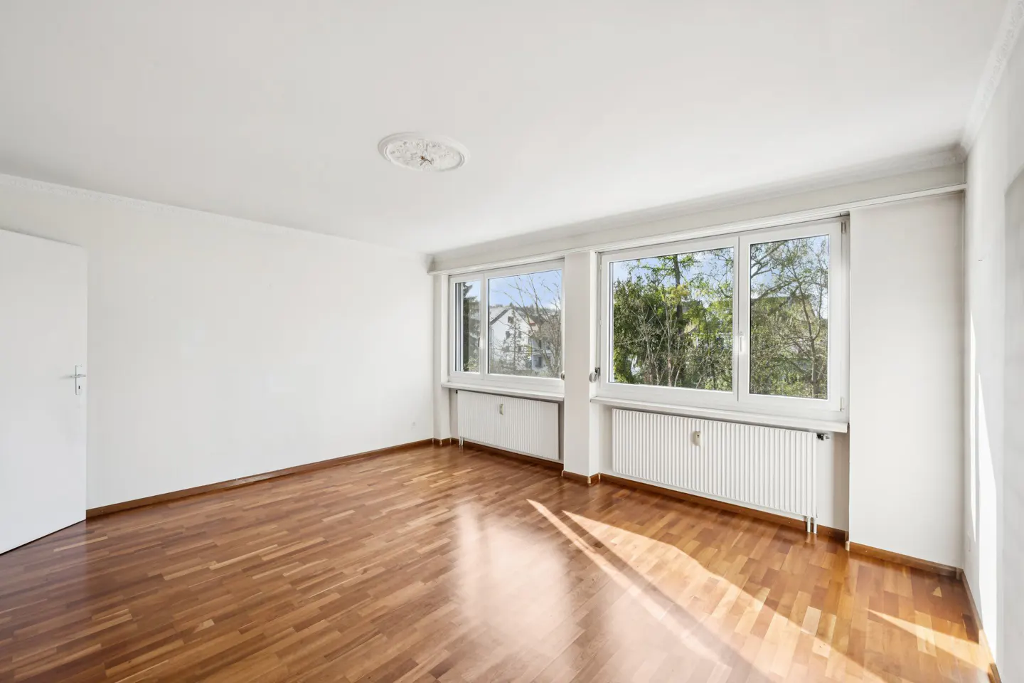 Bright, empty room with hardwood floors, white walls, and two windows showing trees and a building outside.