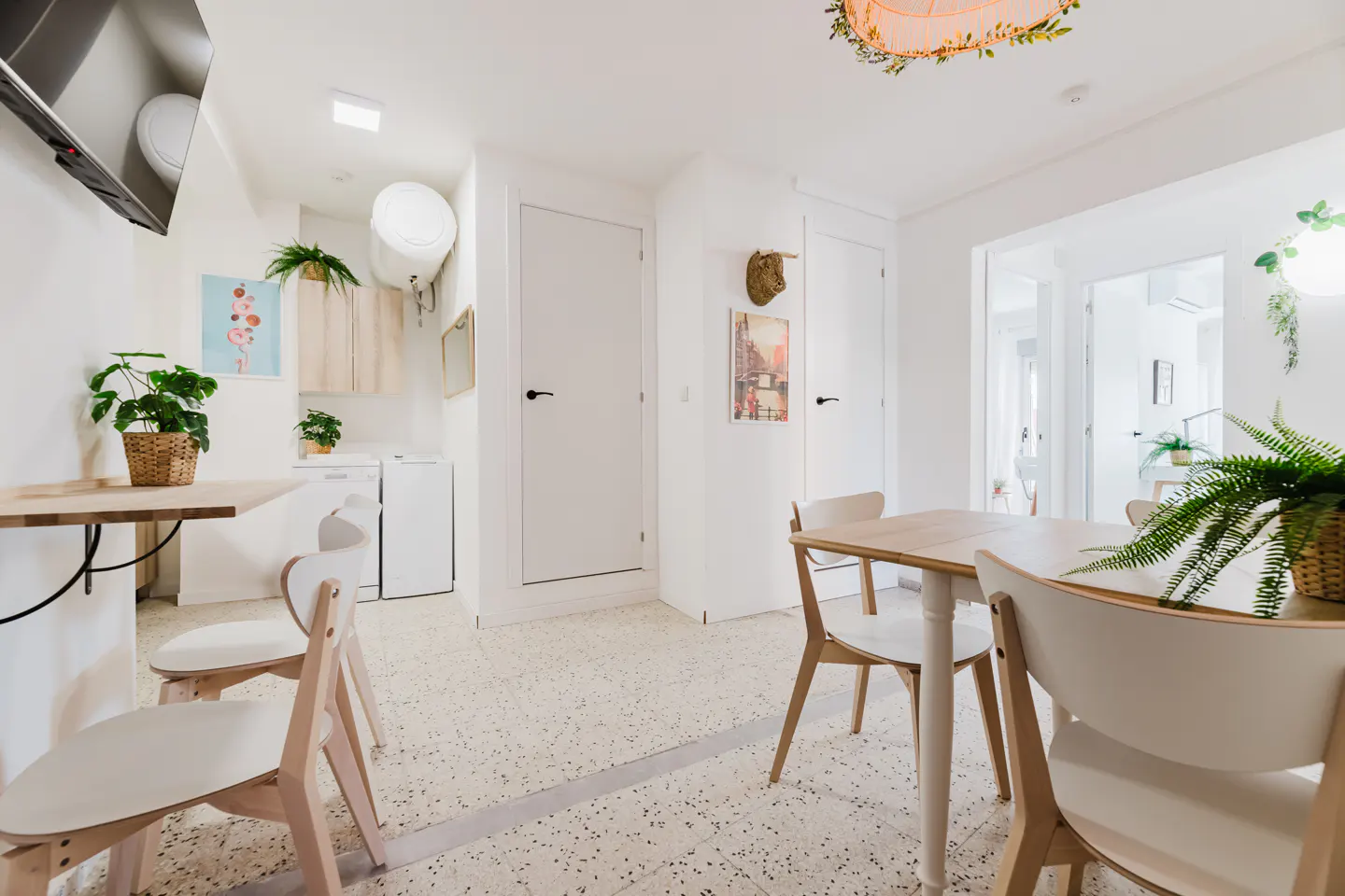 Bright, modern dining area with a light wood table and white chairs. Plants add a touch of nature to the white walls and speckled floor.