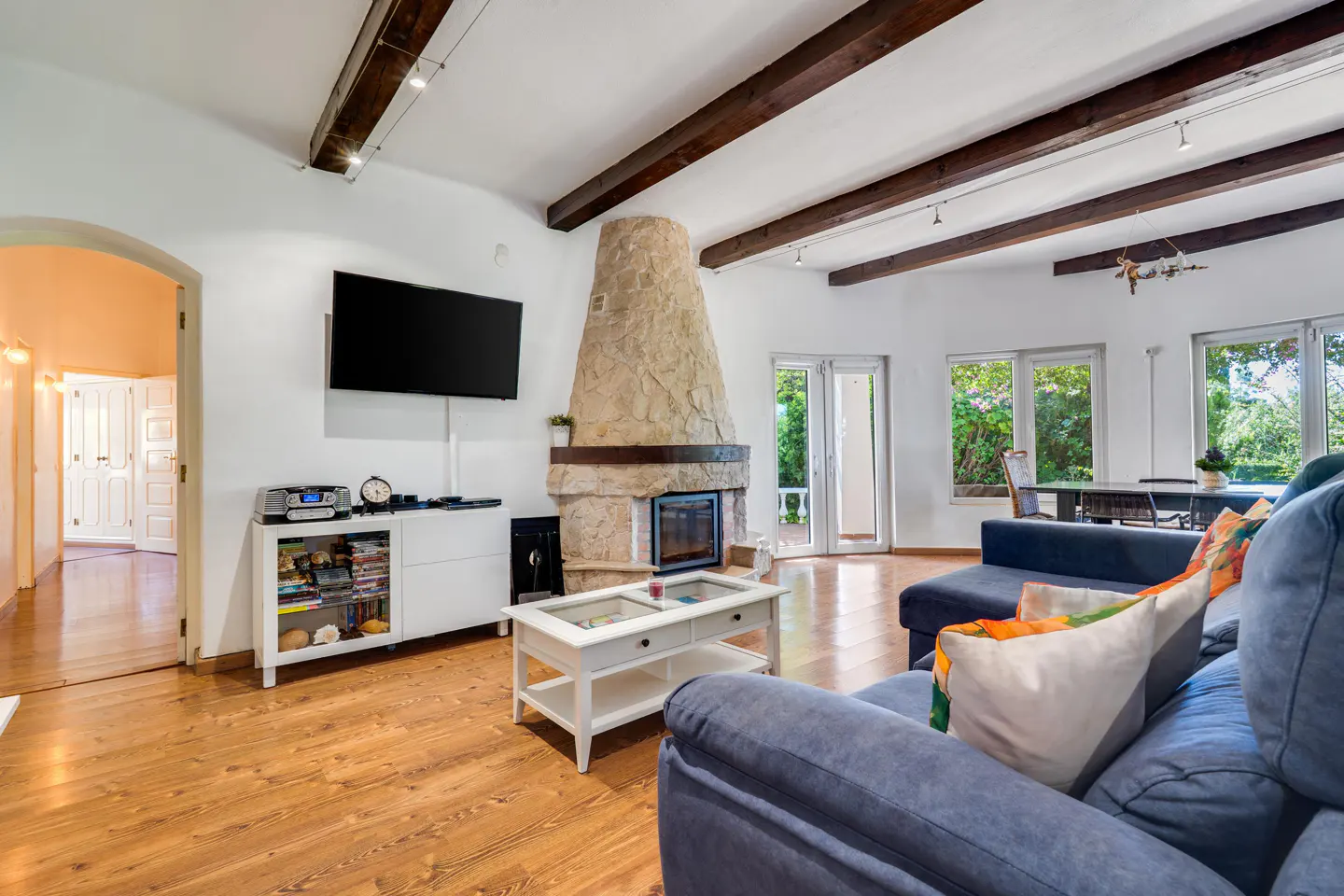 Bright living room with wood floors, white walls, and exposed beams. A stone fireplace, blue sofa, and TV create a cozy space.